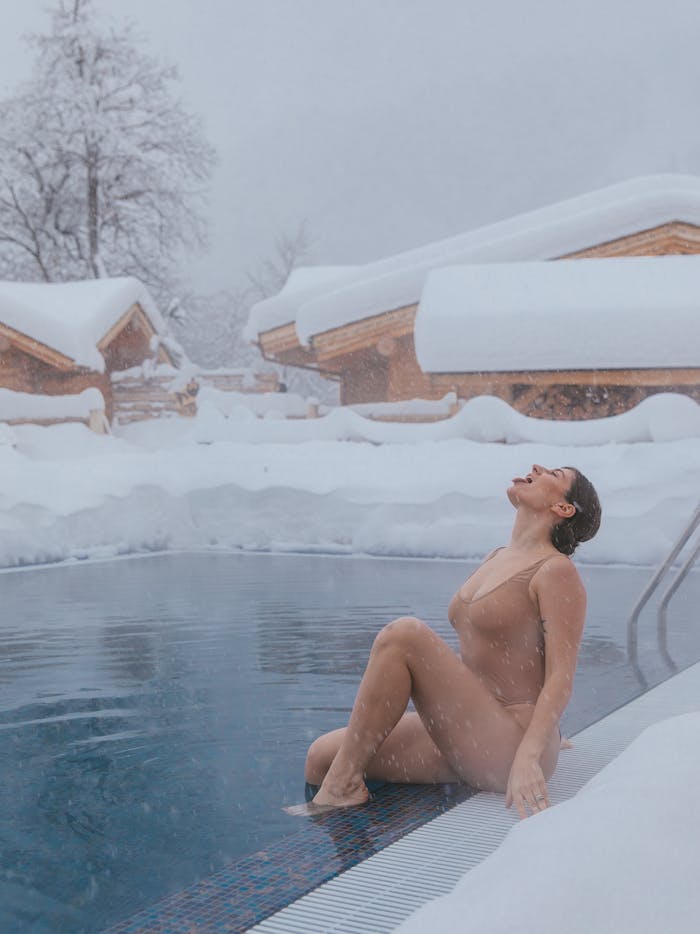 A woman relaxes by an outdoor pool surrounded by snow-covered cabins during winter snowfall.